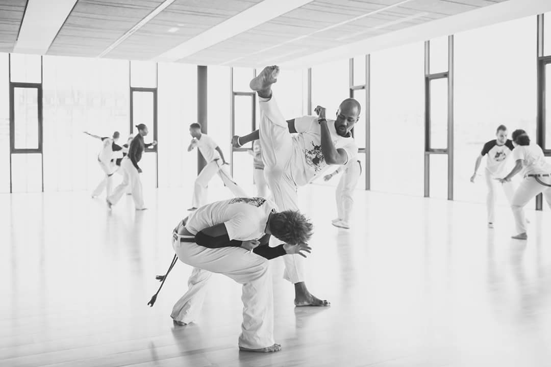 Deux capoeiristes en plein jeu dans une grande salle lumineuse, l'un en coup de pied haut, l'autre en esquive basse.